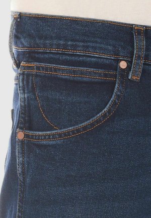 Jeans en denim bleu foncé avec une poche à pièce à l'avant, des accents en métal cuivr�é et des détails de surfilage orange contrastants le long des coutures.