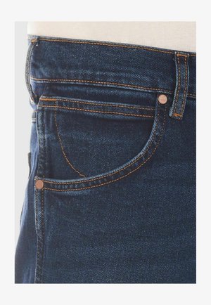 Donkerblauwe denim jeans met een voorzakje voor munten, koperen hardware-accenten en contrasterende oranje stiksel details langs de naden.