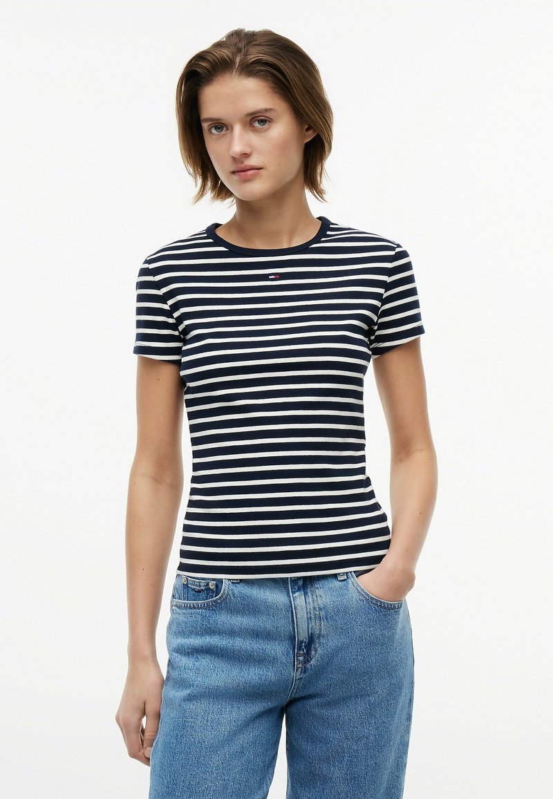 Jeune femme aux cheveux courts portant un t-shirt rayé bleu marine et blanc et un jean bleu clair, debout avec une main dans la poche.