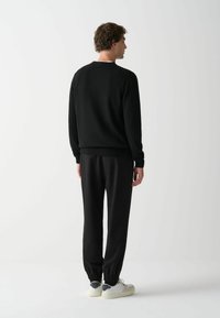 Schwarzer Strickpullover mit einem gerippten Saum, kombiniert mit schwarzer, taillierter Hose und hellen Sneakers mit grauen Akzenten. Rückansicht.
