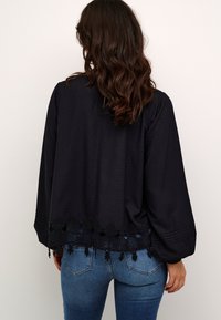 Melnā blūze ar teksturētu audumu, garām piedurknēm un mežģīņu apdari ap apakšmalu. Nedaudz brīvs piegriezums, valkāta pār ziliem džinsiem.