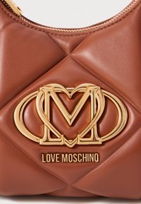Sac à main en faux cuir matelassé marron avec un logo et des éléments en métal doré bien en vue. Texture lisse avec une fermeture zippée sur le dessus.