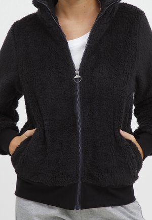 Persona que lleva una chaqueta negra con cremallera y textura peluda, con las manos en los bolsillos laterales y una camisa blanca debajo.