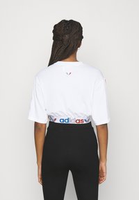 Bílá krátká tričko s krátkým rukávem a kulatým výstřihem, s malým logem na zadní straně a pružným pasem s barevným značením "adidas".