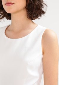 Femme aux cheveux courts bouclés bruns portant un haut blanc sans manches texturé, montrée des épaules au menton sur un fond uni.