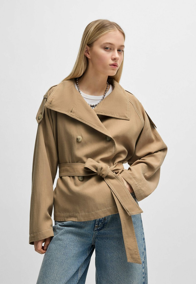 Beige double-breasted jacket with a wide collar, belted waist, and long sleeves. Made from textured fabric, featuring button accents.