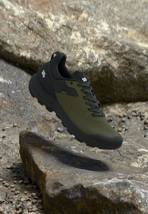 Chaussure de randonnée vert foncé et noir avec une semelle épaisse et des lacets, de la marque "Hanwag", suspendue au-dessus d’un terrain rocheux en plein air.
