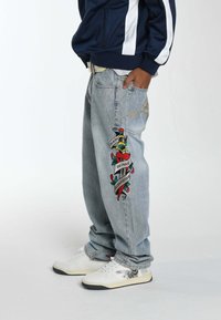 Lichtblauwe, versleten denim jeans met een grote kleurrijke geborduurde ontwerp met een dolk en tekst op het linkerbeen. Witte sneakers worden gedragen.