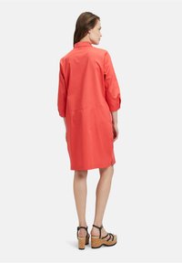 Korallfarbendes Baumwollkleid mit Dreiviertelärmeln und Kragen, mit geradem Schnitt und seitlichen Schlitzen für einen angenehmen Bewegungsfreiheit. Schwarze Riemchensandalen.