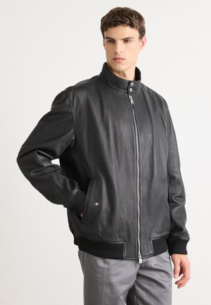 Un jeune homme portant une veste en cuir noir à zip avec col pressionné et poches latérales, associé à un pantalon gris, se tenant contre un fond blanc.