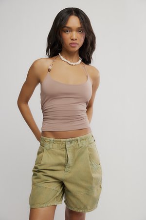 Žena má na sebe taupe halter top, náhrdelník z mušlí a olivovo zelené cargo šortky, stojí pred jednoduchým svetlým pozadím.