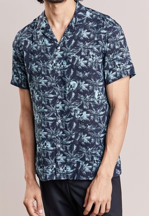 Homme portant une chemise bleu foncé à manches courtes, boutonnée, avec un motif de feuilles de palmier et de crânes bleu clair, associée à un pantalon noir.