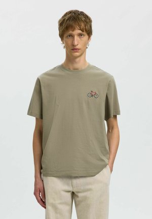 Noor mees kannab helepruuni t-särki, millel vasakul rinnal on väike punane ja sinine jalgrattakujundus, sobitatud beežide pükstega.