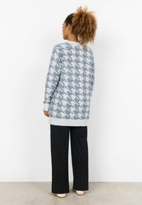 Maglione grigio a motivo houndstooth con una vestibilità più lunga, dettagli a coste nei polsini e nell'orlo, abbinato a pantaloni neri a gamba larga e scarpe neutre.