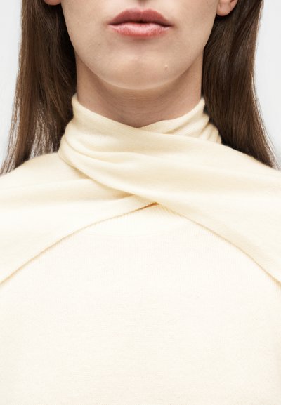 Κοντινό πλάνο του κάτω μέρους του προσώπου και του λαιμού ενός ατόμου που φοράει ένα πουλόβερ κρεμ χρώματος με ένα αντίστοιχο κασκόλ τυλιγμένο γύρω από το λαιμό.
