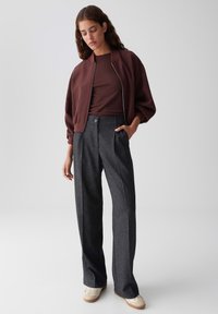 Femme portant une veste bomber bordeaux, un haut en maille marron, un pantalon large gris charbon et des baskets beiges, se tenant avec une main dans la poche.