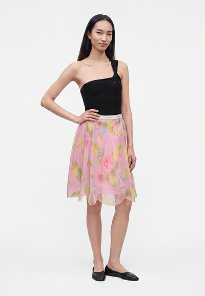 Femme debout dans un haut noir à une épaule, une jupe rose à fleurs longueur genou et des ballerines noires sur un fond blanc uni.