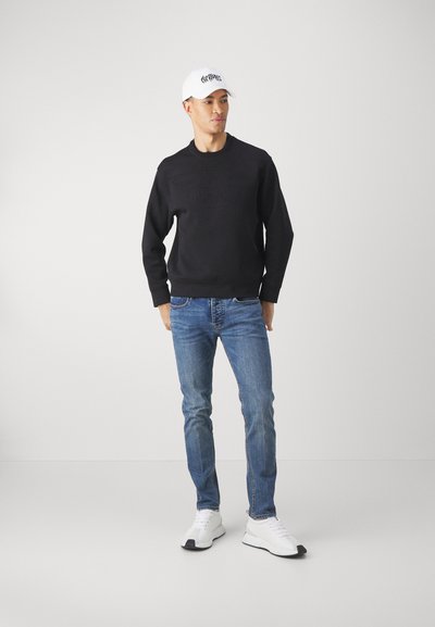 Felpa nera con logo ricamato, jeans blu e sneakers bianchi. Il cappellino è bianco con scritte nere. Il modello si trova su uno sfondo neutro.