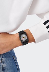 Montre carrée avec un strap en silicone noir, un cadran argenté arborant un point central jaune et des accents colorés (rouge, bleu) avec un design élégant.