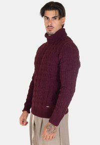 Maglione a coste bordeaux con colletto alto, polsini e orlo a costine. Presenta un motivo testurizzato e una piccola etichetta con logo nella parte inferiore.