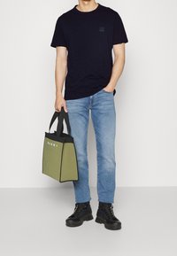 Navy cotton t-shirt, light blue jeans, black lace-up boots. Holding an olive green tote bag with black trim and white logo.