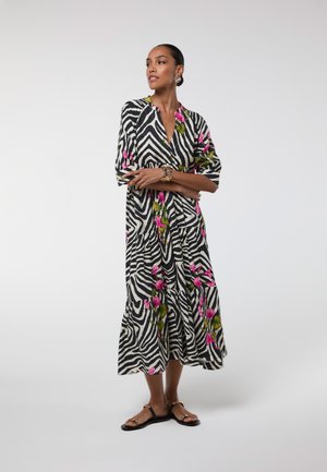 Mujer con un vestido a media pierna en blanco y negro con rayas de cebra y estampados florales rosas, sandalias negras planas y accesorios dorados.