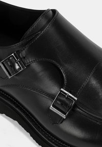 Black leather shoe featuring dual silver buckles, smooth texture, rounded shape, and stitched detailing on the sole.