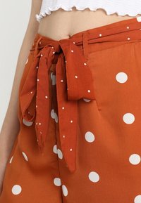 Pantaloni color ruggine con pois bianchi, caratterizzati da una vita legata, una texture liscia e una vestibilità rilassata.
