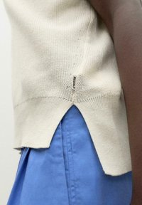 Beige knitted top with a split hem, featuring a small Ecoalf tag on the side. Paired with a blue garment underneath.