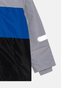 Kleurengeblokt jack met grijze, blauwe en zwarte panelen, gemaakt van glad, lichtgewicht materiaal met reflecterende stripdetails op de mouw.