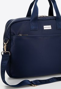 Granatowa torba na laptopa z nylonu z podwójnymi uchwytami, regulowanym paskiem, przednią kieszenią na zamek błyskawiczny oraz metalowym logo. Gładka, lekka tekstura.