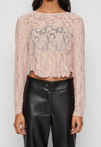 Långärmad, kort topp i genomskinlig ljusrosa blommig spets med scallopade kanter. Matchas med högmidjade svarta läderbyxor med knapprivna detaljer.