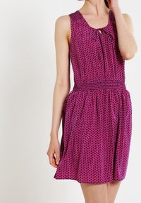 Vestido sin mangas, hasta la rodilla, en color púrpura con un patrón geométrico. Presenta una cintura y un detalle de lazo en el escote.