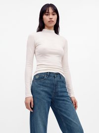 Giovane donna con capelli lisci e scuri indossa una maglia bianca aderente a maniche lunghe e jeans blu a vita alta su uno sfondo semplice.