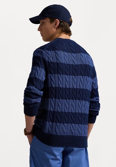 Navyblå kabelstrik sweater med skiftende striber af mørk og lys blå. Ribbet manchetter og talje, båret over lyseblå bukser.