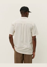 Chemise à manches courtes, couleur beige clair, avec un motif texturé. Dotée d'un empiècement au dos et d'un ourlet arrondi. Associée à un pantalon marron ample.