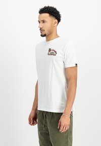 Hvid bomuld t-shirt med kort ærmer, prydet med farverig broderet tekst og palme-træ design på brystet. Bæres med olivenfarvede bukser.