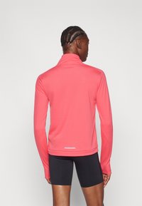 Lagani top s dugim rukavima u svijetloj koraljnoj boji, s visokim ovratnikom i malim bijelim akcentom na rubu. Glatka tekstura, atletski kroj.