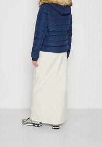 Granatowa kurtka puchowa z futrzanym kapturem, o prążkowanej fakturze i bocznych kieszeniach, zestawiona z długą kremową spódnicą i kolorowymi sneakersami.