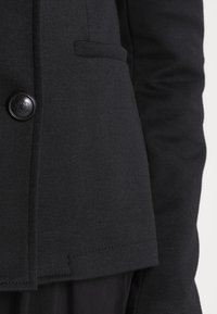 Musta räätälöity bleiseri, joka on valmistettu teksturoidusta kankaasta. Siinä on yksi pyöreä nappi ja sivutasku. Saumadetail näkyy helmassa.