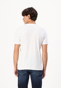 Tricou alb cu mâneci scurte, cu guler rotund, având o textură netedă a materialului, o formă ajustată și un logo mic pe spate, aproape de tiv.