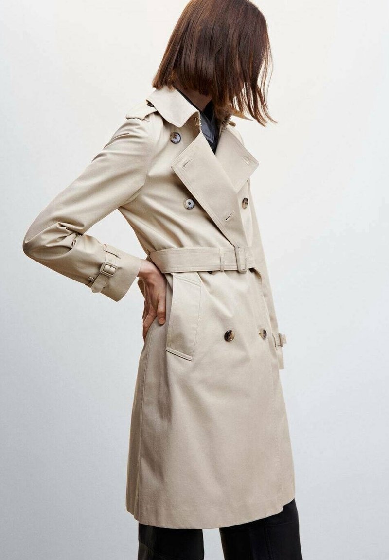 Beżowy trench z podwójnym zapięciem, szerokim kołnierzem i regulowanymi paskami przy mankietach. Posiada paski w talii i boczne kieszenie.