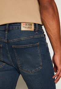 Kék farmernadrág, amely illeszkedő fazonnal, látható varrással és a deréknál elhelyezett "DENIM PROJECT" feliratú bőr címkével rendelkezik.