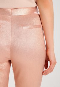 Pantalons ajustés métalliques roses avec une texture brillante, comportant une seule poche arrière et une taille lisse. Main placée sur la hanche.