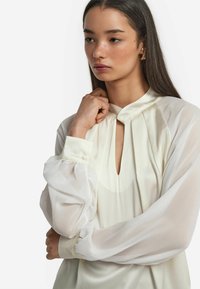 Weiße Bluse mit Schlüsselloch-Ausschnitt und eingeplisster Details, ausgestattet mit transparenten Puffärmeln und satinierte Manschetten mit Knopfakzenten.