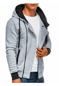 Veste à capuche zippée grise avec une capuche doublée de noir, des poches latérales et des poignets contrastants foncés. Texture douce et coupe ajustée.