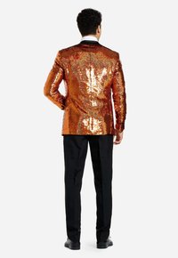 Vista posterior de un hombre con un blazer de lentejuelas naranjas y solapa negra, combinado con pantalones y zapatos negros, mostrando una textura brillante.