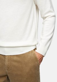 White knitted sweater with ribbed cuffs paired with brown corduroy pants. Sweater features a simple, textured surface and relaxed fit.