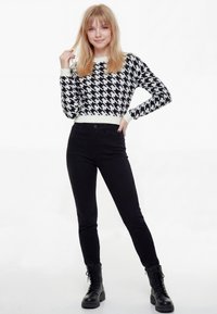Mladá žena s blond vlasmi, oblečená v čierno-bielom svetri s vzorom psieho zubu, čiernych nohaviciach a čiernych čižmách na šnurovanie, stojaca s rukou na bokoch.