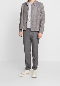 Muž v šedém kostkovaném sáčku, světle šedém tričku s melírem, šedých kalhotách a bílých vysokých teniskách stojící na bílé podlaze.